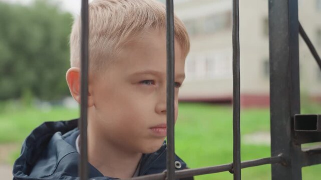 Boy at school gate preparing back to school, somber expression, closeup through metal fence bars, urban campus and green lawn in background, jacket and backpack straps visible, overcast morning light,