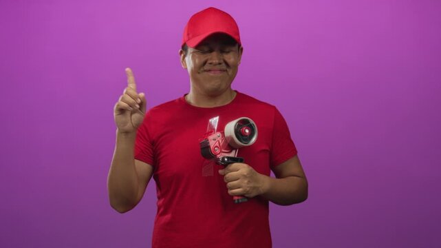 Man in red cap and red shirt holding a packing tape dispenser and pointing index finger at it in a purple studio; duty.