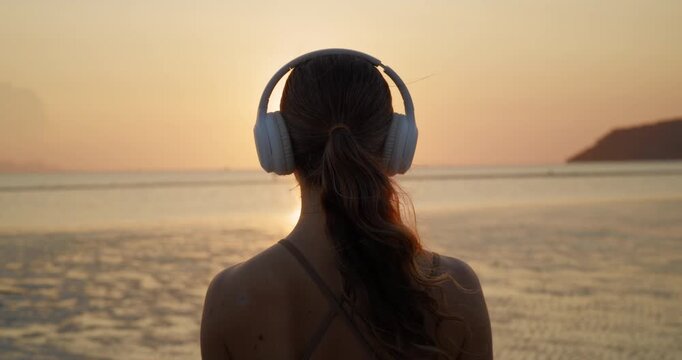 Young woman listens to music via earphones as stands on calm sunset ocean beach backside view. Dreaming female contemplates endless sea surface from coast