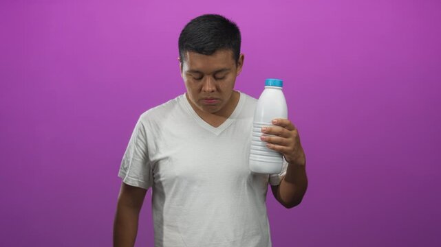 Man wearing white t shirt holding white bottle by cap and pinching fingers in purple studio; thirst refreshment tasting.
