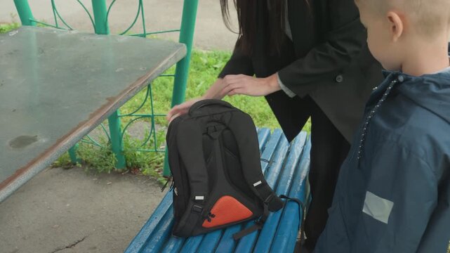 Black teacher checks backpack before school, inspects zipper and straps, arranges notebooks inside bag while white boy watches, park shelter setting, calm guidance and attentive gesture