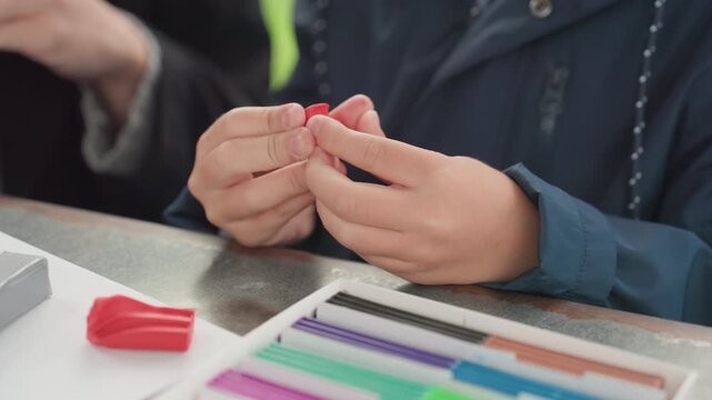 Child hands preparing red eraser on desk with colorful pencils and organizer, closeup of fingers molding small eraser piece, calm focused back to school prep atmosphere, gentle instructional moment