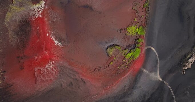 Top-down aerial of a red crater bowl with neon moss and a winding trail through dark ash, creating a striking abstract landform for adventure, mapping, and design visuals.