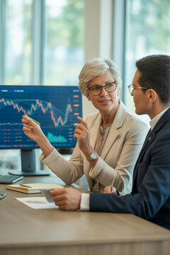 Confident mature senior businesswoman discussing financial charts with a younger male colleague in a modern office