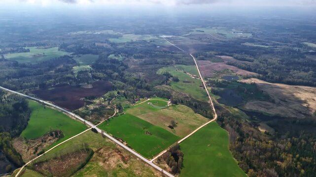 Aerial view of patchwork farmland with crossroads, fields and forest edge under soft daylight. Rural landscape scene with open terrain, winding roads and agricultural land patterns.