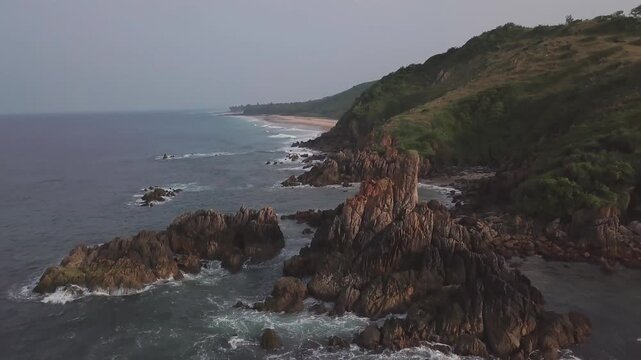 Drone reveals Bhogave Beach with jagged rock formations, coastal vegetation, and turquoise waters along the Arabian Sea coastline in Malvan, Maharashtra, India during overcast afternoon.