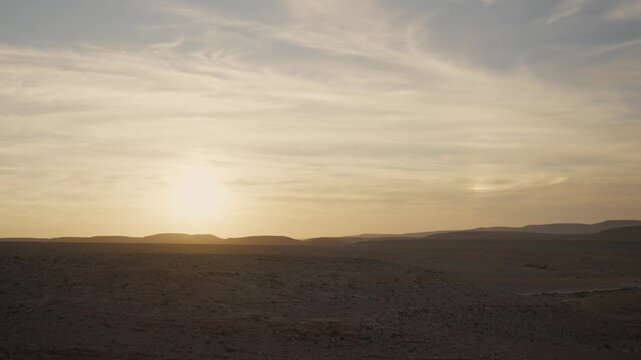 A serene desert landscape on a clear day at sunset or sunrise, sun setting behind distant hills. A peaceful and calm natural environment of blue skies with soft clouds above a wilderness land of dunes