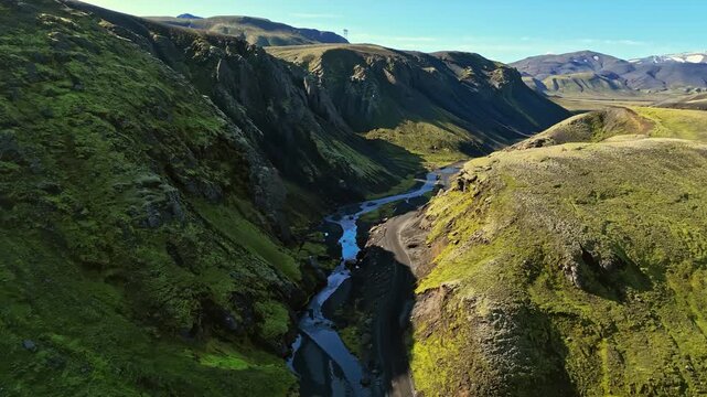 Drone flies through a mossy Iceland canyon above a braided river and gravel road, revealing rugged highlands and mountains under a clear summer sky.