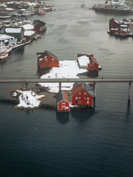 Aerial view of vibrant red cabins nestled on snowy islets, linked by a slender bridge over the dark waters, contrasting against the distant town, Svolvaer, Nordland, Norway.