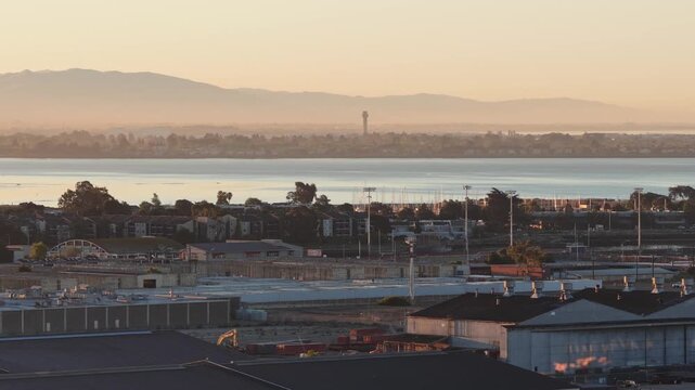 Golden hour light illuminating the industrial buildings and waterfront of Alameda, California, with hazy mountains and the San Leandro bay visible in the warm, atmospheric background