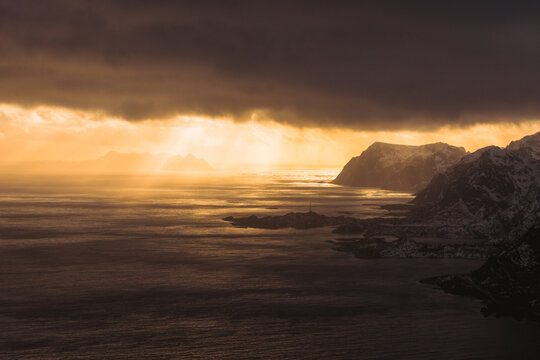 Aerial view of sun rays pierce through dark clouds, illuminating the rugged coastline and tranquil waters of Reinebringen, Nordland, Norway.
