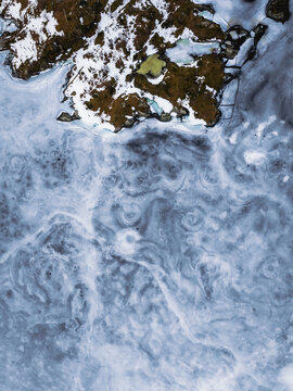 Aerial view of the stark contrast between snow-dusted land and swirling icy waters creates a mesmerizing dance of textures and tones, Tind, Nordland, Norway.