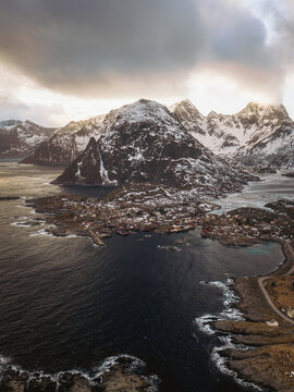 Aerial view of a quaint coastal village nestled beneath snow-dusted peaks, where the dark sea meets the rugged shoreline under a dramatic sky, Tind, Nordland, Norway.