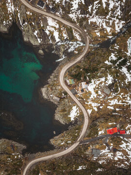 Aerial view of a winding road snaking along the rugged coastline, where turquoise waters meet snow-dusted rocks and quaint buildings, Tind, Norway.