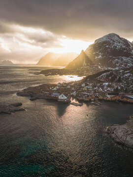 Aerial view of golden sunlight piercing through dramatic clouds, illuminating the rugged coastline and quaint village nestled below snow-dusted mountains, Tind, Nordland, Norway.