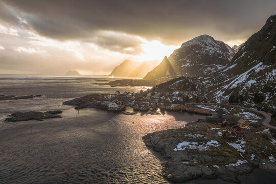 Aerial view of golden sunlight piercing through dramatic clouds over the Lofoten Islands, casting a warm glow on the snow-dusted mountains and the tranquil waters surrounding Tind, Nordland, Norway.