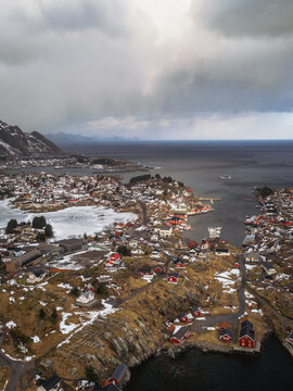 Aerial view of the village nestled between the jagged coastline and the icy waters, a tapestry of red roofs against the snow-dusted landscape, Tind, Nordland, Norway.