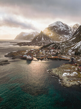 Aerial view of sunlight piercing through the clouds, illuminating the snow-dusted mountains and the tranquil waters surrounding the village, Tind, Nordland, Norway.