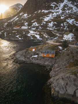 Aerial view of quaint orange cabins nestled on rocky outcrops where land meets the dark, tranquil sea beneath snow-dusted mountains, Tind, Nordland, Norway.