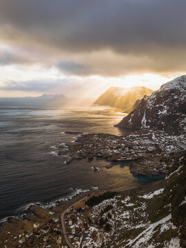 Aerial view of sun rays pierce through dramatic clouds over the snow-dusted mountains and the tranquil waters of the Lofoten Islands, Tind, Nordland, Norway.
