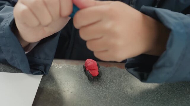 Focused child hands shaping red clay. Outdoor bench craft session showing small eraser on metal desk beside paper and pencil, closeup on fingers molding blue tool, slow methodical motion, calm park