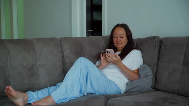 A middle aged woman with long dark hair relaxes on a gray couch in her living room. She is browsing, scrolling, and texting on her smartphone, smiling as she looks at the screen