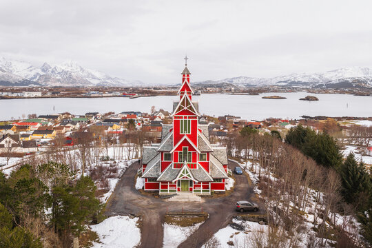 Aerial view of the vibrant red Buksnes Church stands out against the muted winter landscape, with snow-capped mountains in the distance, Buksnes Kirke, Nordland, Norway.