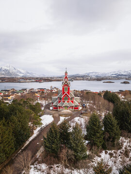 Aerial view of a striking red church standing proudly amidst the green pines and lingering snow, with the serene waters and distant mountains painting a vivid backdrop, Gravdal, Nordland, Norway.