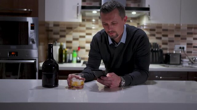 Man Using Smartphone While Drinking Whiskey in Modern Kitchen. A man in a sweater sitting at a kitchen island, scrolling through his phone with a glass of whiskey. Dark bottle on the table