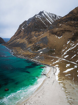 Aerial view of turquoise waters meeting the sandy shore beneath the rugged, snow-dusted peaks of Uttakleivveien, creating a dramatic contrast, Haukland Beach, Nordland, Norway.