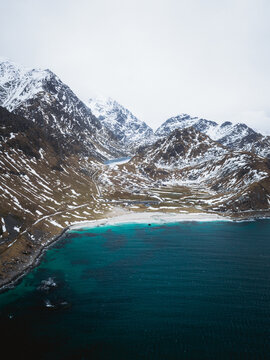 Aerial view of turquoise waters meeting snow-dusted mountains, a serene contrast of warmth and chill at Haukland Beach, Leknes, Nordland, Norway.