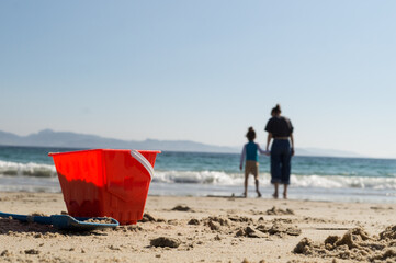 Ground-level view on a sandy beach, with a bright red toy bucket and blue shovel in sharp focus in the foreground. In the blurry background, a child and a mother walk hand-in-hand towards the ocean un © Phoscar