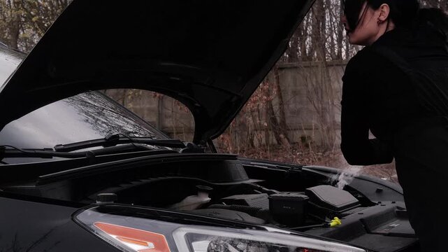female mechanic in work uniform opens hood of car with smoke coming out. emergency stop, repair, problem, black modern car, service, under hood, wrench