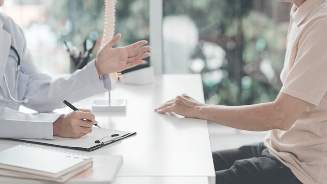 The doctor is advising an elderly patient and explaining the anatomy using an anatomical model of the human pelvis and spine in the examination room.