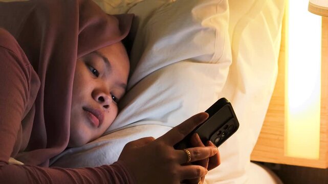 Insomnia and sleep problems. An Asian Muslim woman with smartphone in her hands lying awake on bed at night.