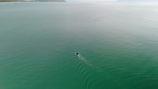 A person rides a small boat on the clear water near DokLet beach in Vietnam. The beach stretches along the coast with green hills in the background and boats nearby