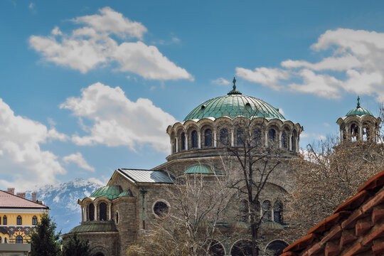 Historic St. Nedelya Cathedral in Sofia, Bulgaria, showcasing its distinctive green copper domes and Neo-Byzantine stone architecture. In the background, the snow-capped peaks of Vitosha Mountain 