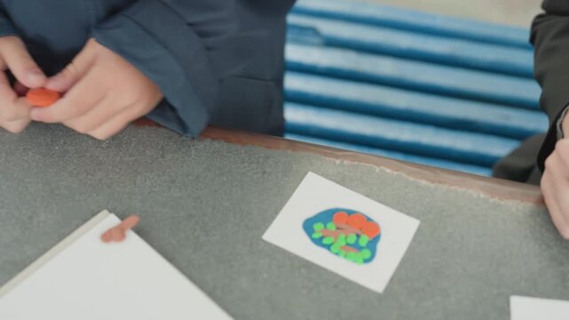 Child hands crafting colorful clay flower on bench, student molding orange petal with fingers, small paper card with bright floral design, jacketed child at school table preparing craft to take back