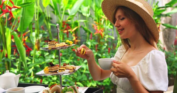 Woman wearing straw hat enjoying afternoon tea in lush tropical garden, holding white cup and reaching for pastry from tiered stand, bright sunny lighting, luxury vacation lifestyle, elegant dining ex