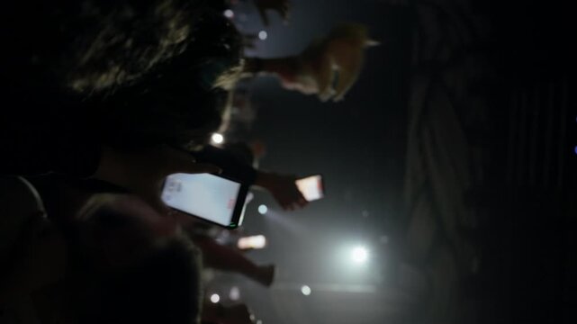 An energetic crowd at a live concert or event captures the performance on their smartphones, with hands raised high and phone screens illuminating the dark venue. Camera rotates from vertical to