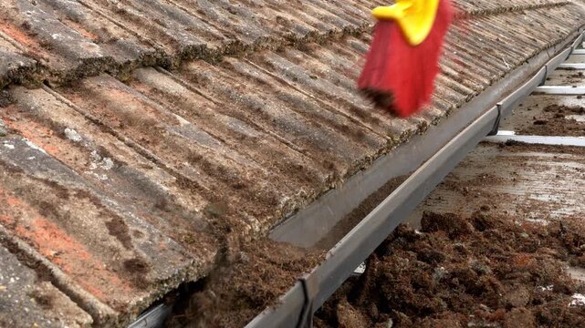 Closeup elevated view of a thin brush on a pole, dragging dried moss along a rain gutter for removal, which has been cleaned off the tiles of a building roof.