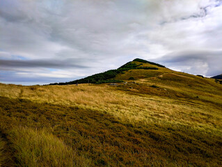 Fototapeta premium Rolling grassy hills with a winding trail leading to a mountain peak beneath a dramatic cloudy sky, ideal for outdoor, adventure, and nature-themed designs.
