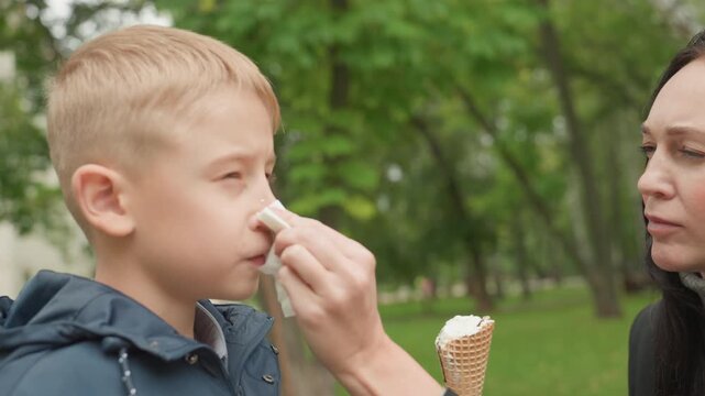 Park tissue wipe, white mother cleans childs nose preparing for back to school. Mother dabs runny nose with napkin while child holds melting cone, calm supportive moment under green trees, close