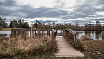 Obraz premium Wooden lakeside viewing platform and boardwalk overlooking calm pond under dramatic cloudy winter sky