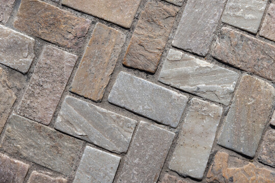 Close-up of textured stone pavers arranged in a herringbone pattern, showing natural color variations, rough surfaces and mortar joints. Background photo texture for architecture and landscaping