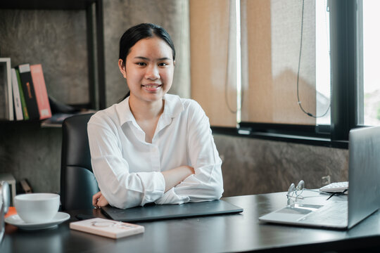 Young woman in a white shirt sitting at a desk in a modern office, smiling confidently with a laptop and documents.