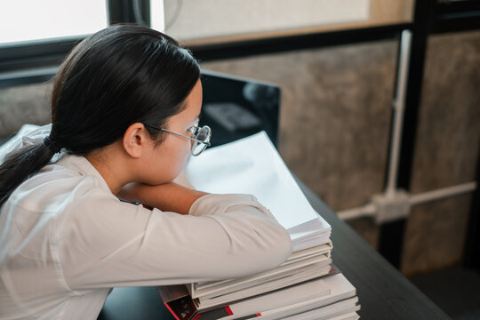 Young woman in glasses leans on books, reflecting in a modern office. Ideal for themes of education, work, or contemplation.