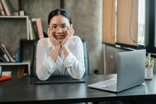 A young woman in a modern office, smiling confidently at her desk with a laptop and books, embodying professionalism and success.