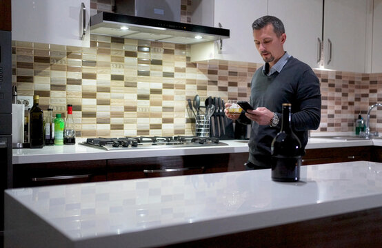 Man Using Smartphone While Drinking Whiskey in Modern Kitchen. A man in a sweater walking at a kitchen island, using through his phone with a glass of whiskey. Dark bottle on the table