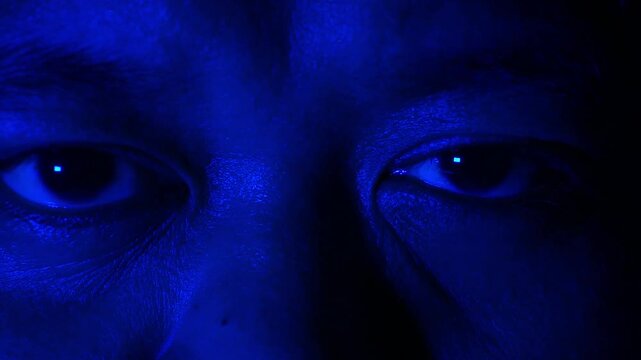 Dynamic shifting colored lights in red, blue, and green wash over a male face, illuminating his intense eyes staring directly ahead in deep shadows.